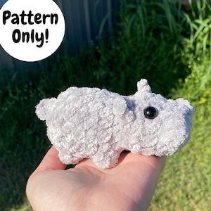 May include: A gray plush amigurumi hippopotamus toy with black eyes. The toy is being held in a person's hand.