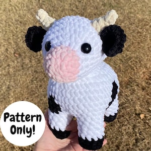 May include: A crocheted plush toy cow with white fur, black spots, and a pink nose. The cow has a white and brown yarn headband and black yarn ears. The toy is being held in a hand, and the text "Pattern Only!" is visible in the background.