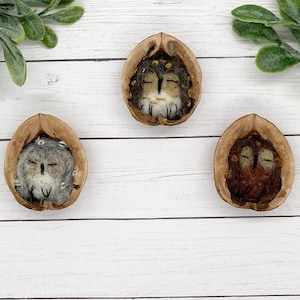 May include: Three handcrafted felted owl figurines nestled in walnut shells. Each owl has closed eyes and is made with brown, gray, and white wool. The shells are arranged on a white wooden surface.