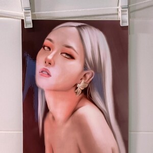 May include: A portrait print of a woman with long, silver hair and a fair complexion. She wears a gold earring and has a slight smile. The print is held by two white clothespins on a white string.