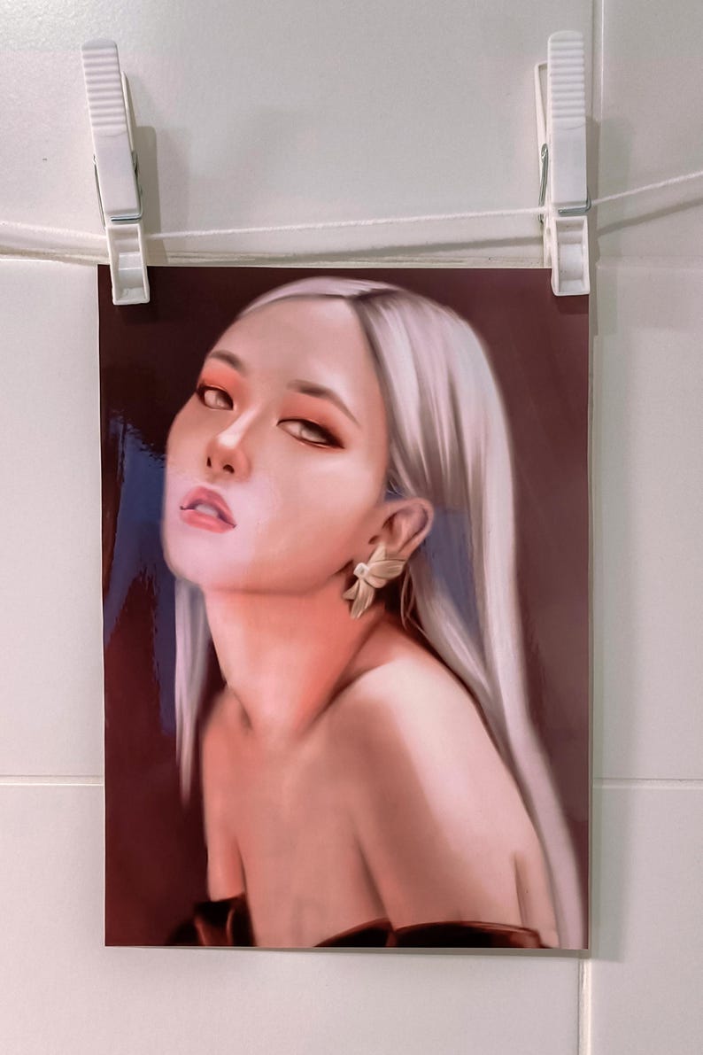 May include: A portrait print of a woman with long, silver hair and a fair complexion. She wears a gold earring and has a slight smile. The print is held by two white clothespins on a white string.