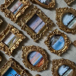 May include: A collection of miniature framed paintings with ornate gold-colored frames. The paintings depict various landscape scenes, including seascapes and sunsets. The frames are in different shapes, including oval, square, and rectangular.