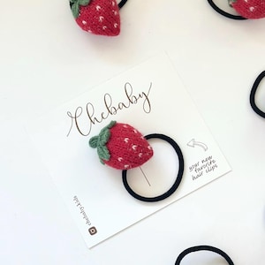 May include: A collection of red and green strawberry-shaped hair ties. The strawberries are knitted with white speckles and green leaves. The hair ties are on a white card with the text "Chebaby" and "your new favorite hair clips".