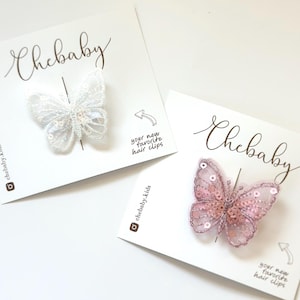 May include: Two butterfly hair clips, one white and one pink, with sequins and lace. The clips are displayed on white cards with the text "Chebaby" and "your new favorite hair clips".