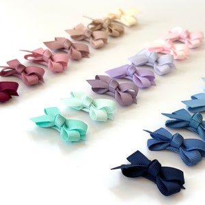 Puede incluir: Un conjunto de 15 mini lazos para el pelo en varios colores, incluyendo burdeos, rosa, lavanda, azul claro y azul marino. Los lazos est&aacute;n dispuestos en un patr&oacute;n de arco&iacute;ris sobre un fondo blanco.