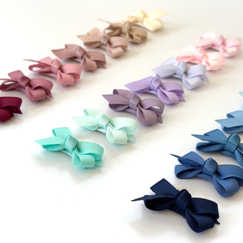 Puede incluir: Un conjunto de 15 mini lazos para el pelo en varios colores, incluyendo burdeos, rosa, lavanda, azul claro y azul marino. Los lazos est&aacute;n dispuestos en un patr&oacute;n de arco&iacute;ris sobre un fondo blanco.