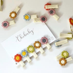 Könnte beinhalten: Eine Sammlung von Haarspangen mit gestickten Blumenmotiven in verschiedenen Farben wie Orange, Gelb, Blau, Grün und Pink, befestigt an cremefarbenen gerippten Stoffspangen, präsentiert mit einer Karte mit der Aufschrift Chebaby.