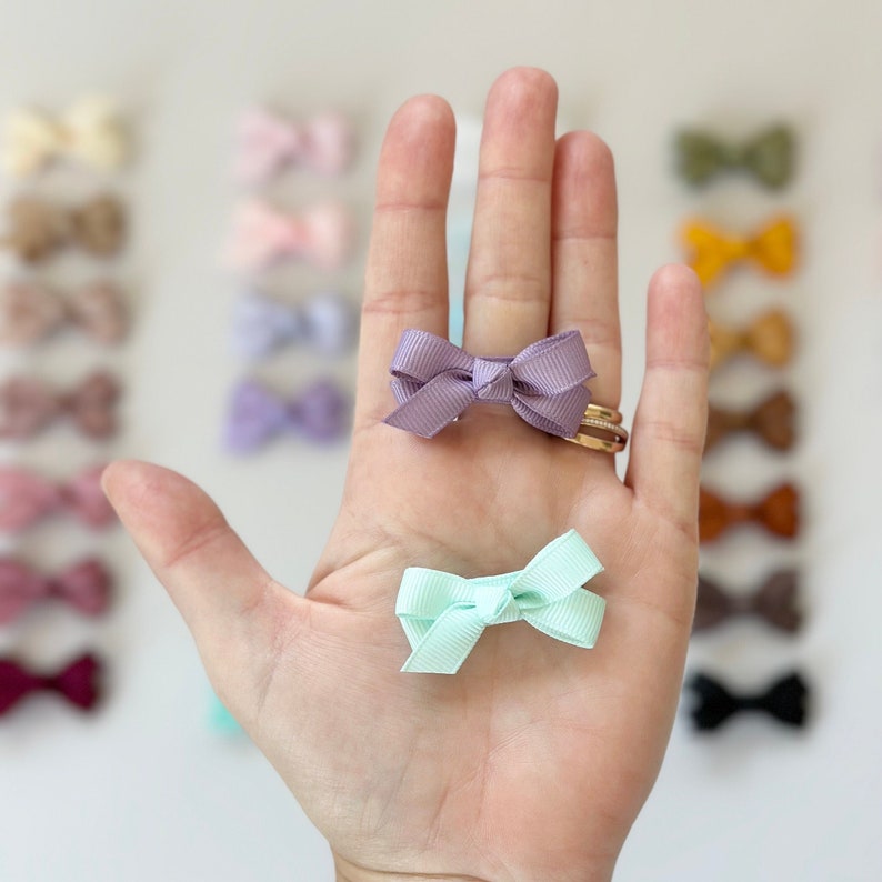 Puede incluir: Dos lazos peque&ntilde;os para el cabello, uno lavanda y otro verde claro, se muestran en una mano. Los lazos est&aacute;n hechos de cinta de sat&eacute;n y est&aacute;n atados en forma de lazo simple.