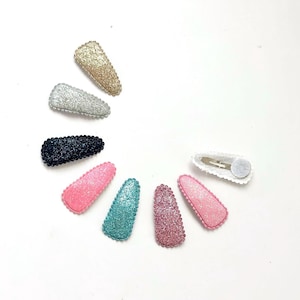 Könnte beinhalten: Eine Sammlung von acht glitzernden Haarspangen in verschiedenen Farben, darunter Gold, Silber, Schwarz, Rosa, Türkis und Lila. Jede Spange hat eine ovale Form mit einer strukturierten, glitzernden Oberfläche und einem Metallverschluss. Die Spangen sind halbkreisförmig angeordnet.