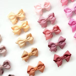 May include: A collection of 14 small, handmade hair bows in various shades of pink, peach, and orange. The bows are made from satin ribbon and are arranged in a gradient pattern.