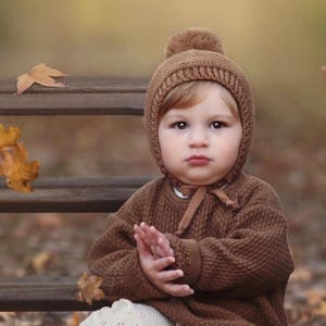May include: A brown knitted sweater and a matching brown knitted bonnet with a pom-pom on top. The bonnet features a ribbed edge and ties under the chin with brown fabric ribbons. The sweater has a textured, waffle-like knit pattern.
