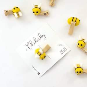 Könnte beinhalten: Eine Sammlung von gelb-schwarzen Filz-Bienen-Haarspangen mit weißen Flügeln und schwarzen Augen. Die Spangen sind an hellbraunen Metallclips befestigt. Eine weiße Karte mit dem Text "Chebaby" ist ebenfalls vorhanden.