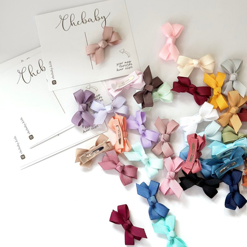 Puede incluir: Una colecci&oacute;n de 20 peque&ntilde;os lazos para el cabello de colores con pinzas. Los lazos est&aacute;n hechos de cinta de sat&eacute;n y vienen en una variedad de colores, incluyendo rosa, azul, morado, verde, amarillo y rojo. Los lazos est&aacute;n dispuestos en un patr&oacute;n aleatorio sobre un fondo blanco. El texto "tus nuevas pinzas para el cabello favoritas" es visible en una tarjeta blanca.