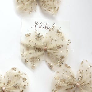 Puede incluir: Varios lazos para el pelo de tul marfil con detalles de purpurina dorada se muestran sobre una superficie blanca. Cada lazo está atado con una cinta rosa pálido. Se ve una tarjeta blanca con el texto "Chebab".