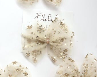 Puede incluir: Varios lazos para el pelo de tul marfil con detalles de purpurina dorada se muestran sobre una superficie blanca. Cada lazo está atado con una cinta rosa pálido. Se ve una tarjeta blanca con el texto "Chebab".