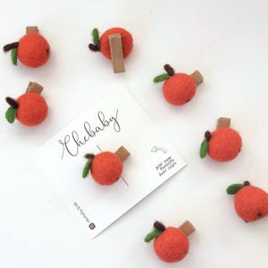 May include: Orange felt apple hair clips with green leaves and brown stems. The clips are arranged around a white card with the text "Chebaby" and "your new favorite hair clips".