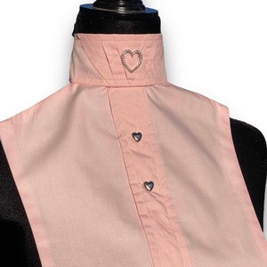 May include: A pink sleeveless shirt with a white collar and a heart-shaped pin. The collar has a pleated design and the shirt has two heart-shaped buttons.