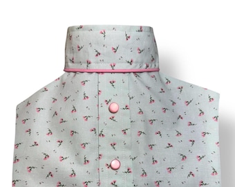 May include: A light blue garment with a floral pattern of small pink flowers. The collar is trimmed with pink piping, and the garment fastens with pink buttons. The fabric has a delicate, feminine aesthetic.