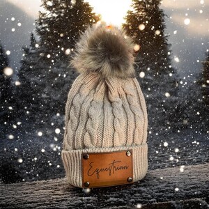 May include: A beige cable-knit beanie with a faux fur pom-pom. The hat features a brown leather patch with the word "Equestrian" in script. The background shows a snowy forest scene.