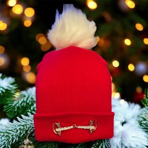 May include: A bright red beanie with a white pom-pom. The hat features a gold-colored equestrian bit design on the brim. The background includes a Christmas tree with blurred lights and snowy branches.