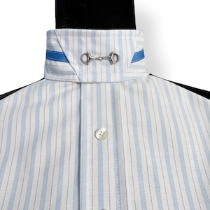 May include: Close-up of a white and light blue striped shirt with a high collar. The collar features a silver bit detail and blue ribbon accents. The shirt has white buttons and a classic design.