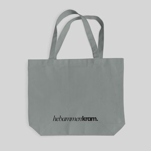 May include: A grey tote bag with two shoulder straps and the black text "hebammenkram." printed in a serif font near the bottom center. The bag features a rectangular body with a flat base and a simple, functional design for carrying items.