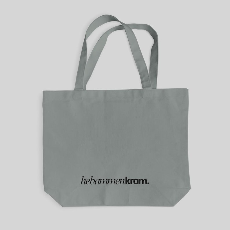 May include: A grey tote bag with two shoulder straps and the black text "hebammenkram." printed in a serif font near the bottom center. The bag features a rectangular body with a flat base and a simple, functional design for carrying items.