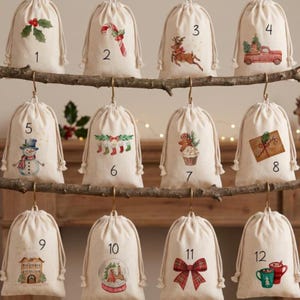 May include: Twelve off-white drawstring canvas bags hanging from a wooden branch. Each bag features a unique Christmas-themed illustration and a black number from 1 to 12, including holly, a candy cane, a reindeer, a truck, a snowman, and stockings.