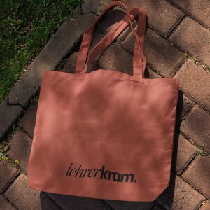 May include: A terracotta-colored tote bag with the text "lehrerkram." printed in black. The bag has two handles and is made of a soft material. The bag is sitting on a brick surface.