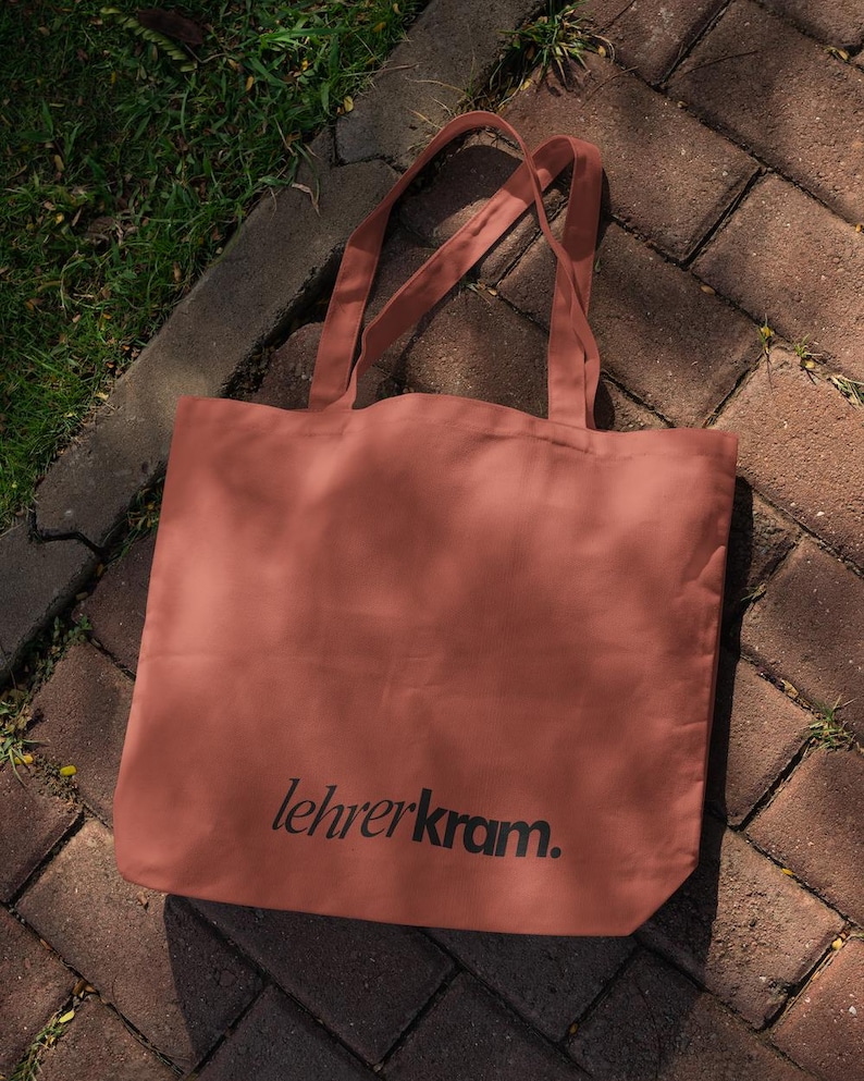 May include: A terracotta-colored tote bag with the text "lehrerkram." printed in black. The bag has two handles and is made of a soft material. The bag is sitting on a brick surface.