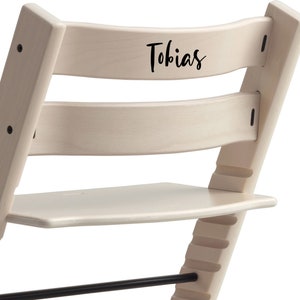 Puede incluir: Una silla alta de madera blanca con un reposapi&eacute;s negro y una placa de nombre negra en la parte posterior que dice "Tobias".