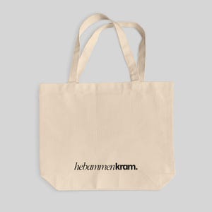 May include: A beige canvas tote bag with two shoulder straps. The bag features the black text "hebammenkram." printed in a serif font on the lower center portion of the front panel. The design is simple, functional, and rectangular in shape.