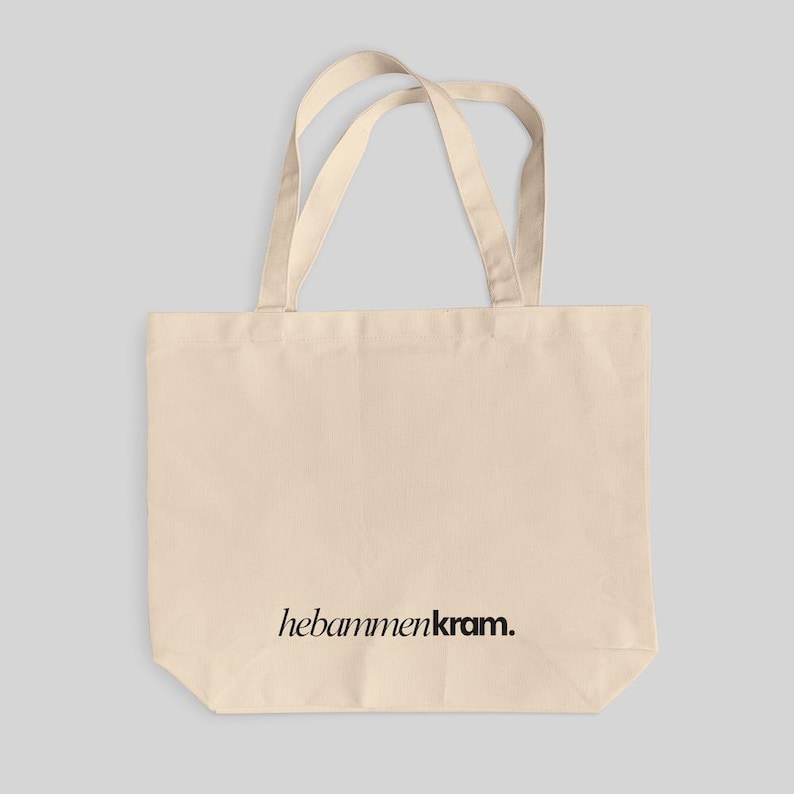 May include: A beige canvas tote bag with two shoulder straps. The bag features the black text "hebammenkram." printed in a serif font on the lower center portion of the front panel. The design is simple, functional, and rectangular in shape.