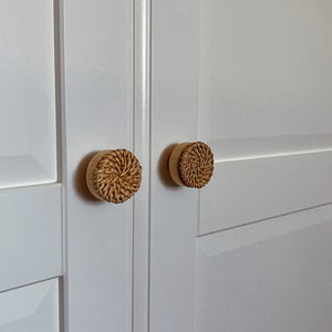以下が含まれることがあります： 白く塗られたキャビネットのドア2枚に、籐で編まれたノブが付いている。ノブは丸く、自然な木の色をしている。