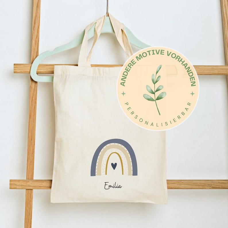 May include: A cream-coloured tote bag featuring a rainbow design in blue, tan, and gold, with a small blue heart. The name "Emilia" is printed below the rainbow. The bag is hanging on a light green hanger.