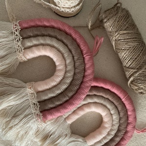 May include: Two pink, brown, and white yarn rainbows with fringe and lace trim. The rainbows are handmade and are a popular home decor item.