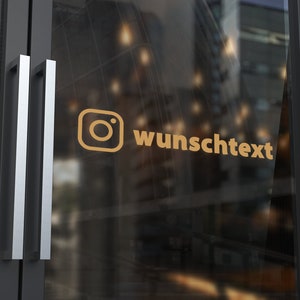 May include: A gold-coloured Instagram logo icon and the text "wunschtext" applied as a decal on a glass door with a dark grey frame. The design is positioned horizontally on the transparent surface, featuring a clean, sans-serif typeface.