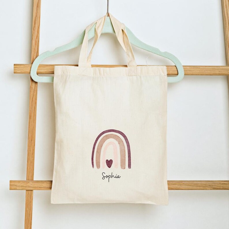 May include: A cream-coloured tote bag featuring a rainbow design in shades of brown and pink, and a heart. The name "Sophia" is written below the rainbow in a script font. The bag hangs on a light green hanger.