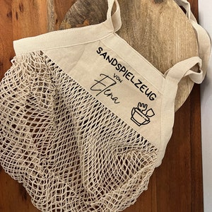 Puede incluir: Una bolsa de malla beige con una parte superior de lona blanca. La parte superior de lona tiene texto negro que dice "Sandspielzeug von Elena" y un icono negro de un cuenco con una pala y un rastrillo.