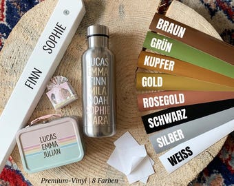 Puede incluir: Una botella de agua de metal plateado con nombres impresos verticalmente, una pequeña lata rectangular con un arcoíris pastel y nombres, una tira de madera blanca con nombres y ocho tiras de vinilo de colores etiquetadas Braun, Grün, Kupfer, Gold, Rosegold, Schwarz, Silber, Weiss.