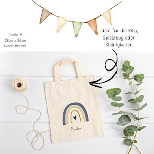 May include: A cream tote bag featuring a blue, tan, and gold rainbow design and the name "Emilia". The bag has a short handle and measures 28cm x 32cm. A string of triangular flags hangs above. Text on the bag says "ideal f&uuml;r die Kita, Spielzeug oder Kleinigkeiten".