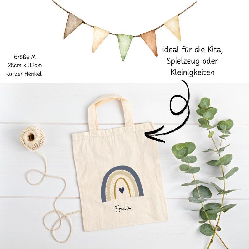May include: A cream tote bag featuring a blue, tan, and gold rainbow design and the name "Emilia". The bag has a short handle and measures 28cm x 32cm. A string of triangular flags hangs above. Text on the bag says "ideal f&uuml;r die Kita, Spielzeug oder Kleinigkeiten".
