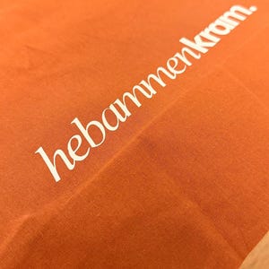 May include: A vibrant orange fabric surface featuring the white text "hebammenkram." printed in a clean, sans-serif font. The material displays a visible woven texture, and the lettering is positioned diagonally across the frame.