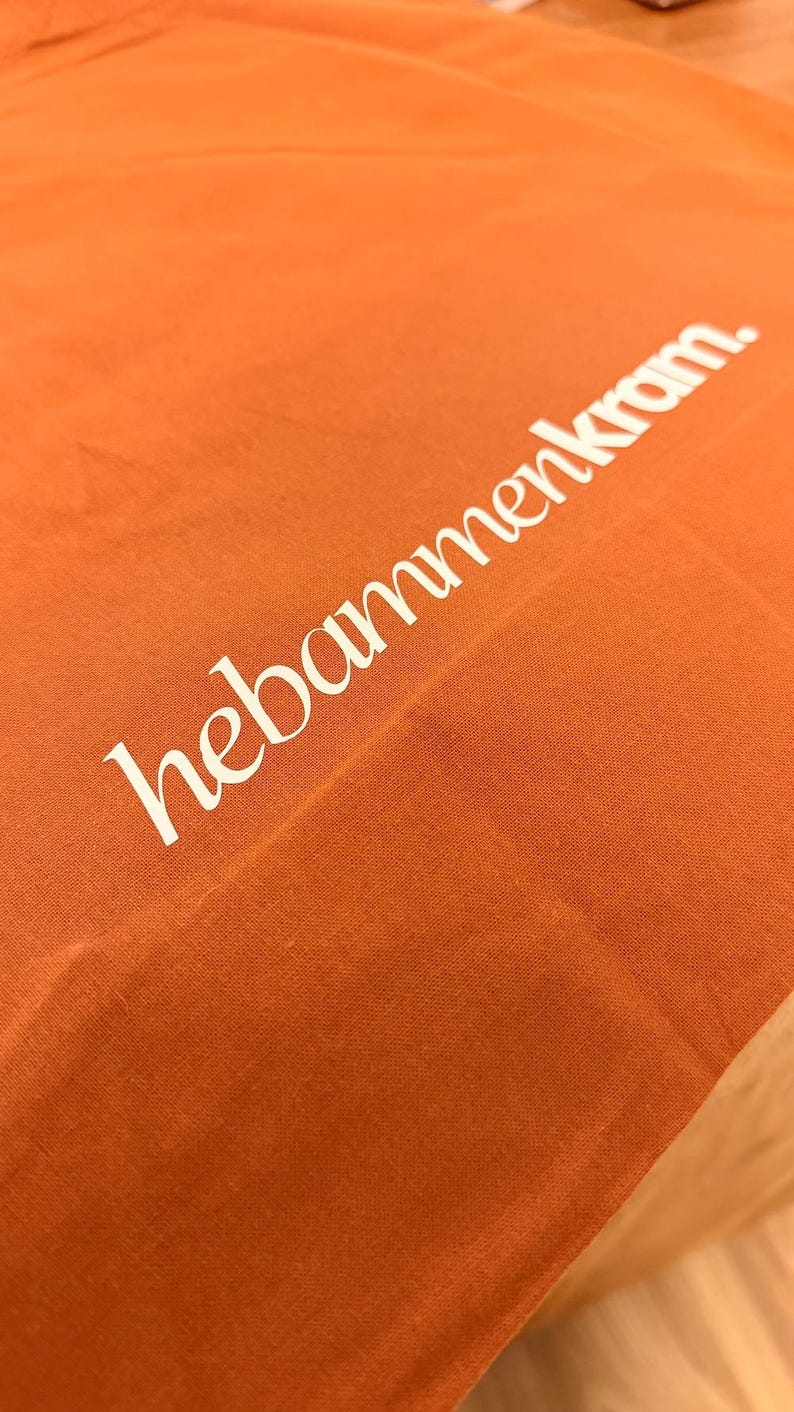 May include: A vibrant orange fabric surface featuring the white text "hebammenkram." printed in a clean, sans-serif font. The material displays a visible woven texture, and the lettering is positioned diagonally across the frame.