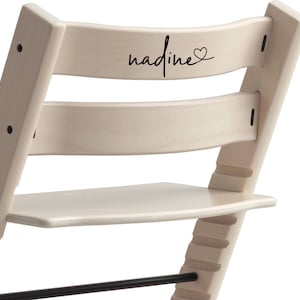 Puede incluir: Una silla de madera blanca con una inscripci&oacute;n negra que dice "nadine" con un s&iacute;mbolo de coraz&oacute;n. La silla tiene un reposapi&eacute;s y una barra de metal negro.
