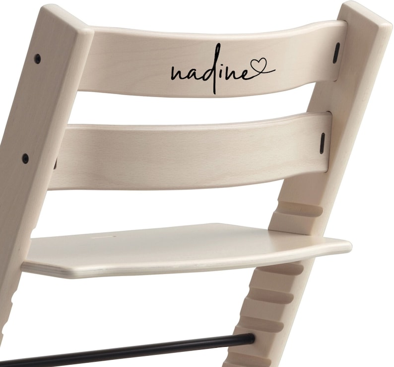 Puede incluir: Una silla de madera blanca con una inscripci&oacute;n negra que dice "nadine" con un s&iacute;mbolo de coraz&oacute;n. La silla tiene un reposapi&eacute;s y una barra de metal negro.
