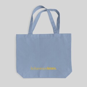 May include: A light blue tote bag with two shoulder straps and the text "hebammenkram." printed in yellow on the lower front section. The bag features a rectangular shape with a flat bottom and is constructed from a solid, opaque fabric material.