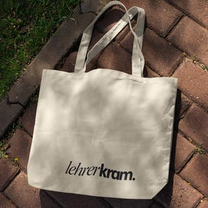 May include: Cream-colored tote bag with the text "lehrerkram." printed in black. The bag has two long handles and is made of a soft, durable fabric. The bag is sitting on a brick surface with grass in the background.