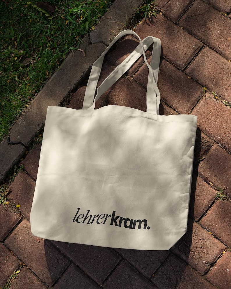 May include: Cream-colored tote bag with the text "lehrerkram." printed in black. The bag has two long handles and is made of a soft, durable fabric. The bag is sitting on a brick surface with grass in the background.