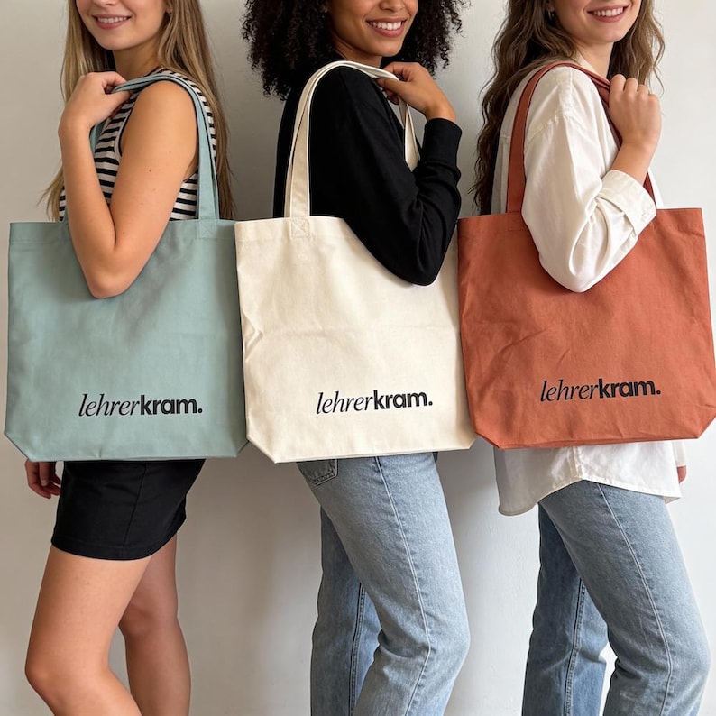 May include: Three tote bags in different colors: light blue, off-white, and rust. Each bag has a rectangular shape with a long handle and the text "lehrerkram." printed in black. The bags are held by three people.